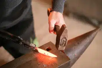 Idée cadeau anniversaire DIY pour son mari, sa femme ou un proche créatif : atelier couteau artisanal Wecandoo avec artisan forgeron, établi, lame en acier en cours de polissage et manche en bois travaillé, ambiance atelier authentique et lumineuse, mettant en valeur la tradition, la patience et la fierté de repartir avec un couteau unique fait main.