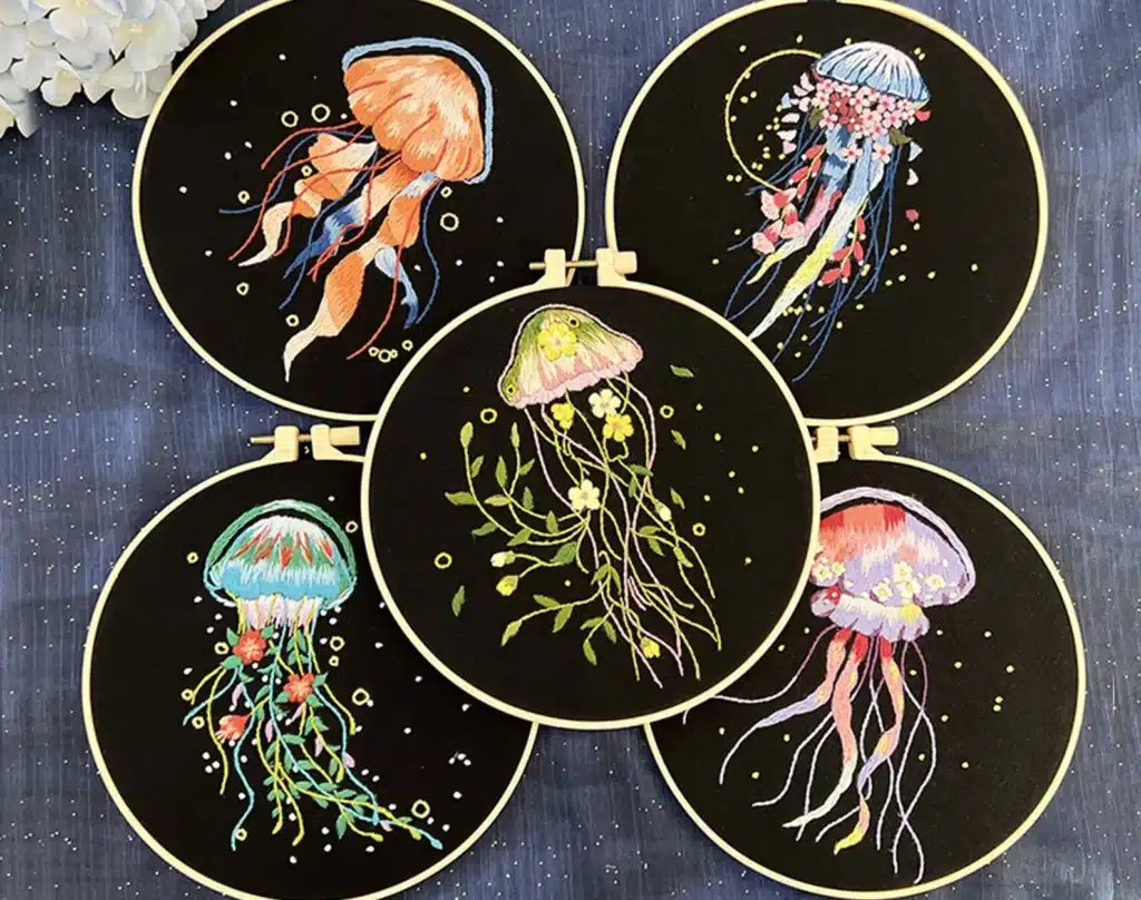 Idée cadeau anniversaire DIY pour sa femme, sa sœur ou son amie créative : kit de broderie Etsy motif méduses et fleurs comprenant toile imprimée, fils colorés, cercle à broder et aiguilles, posé sur une table claire avec une broderie en cours, ambiance artisanale et douce, mettant en valeur la créativité fait main et l’esprit décoratif poétique.