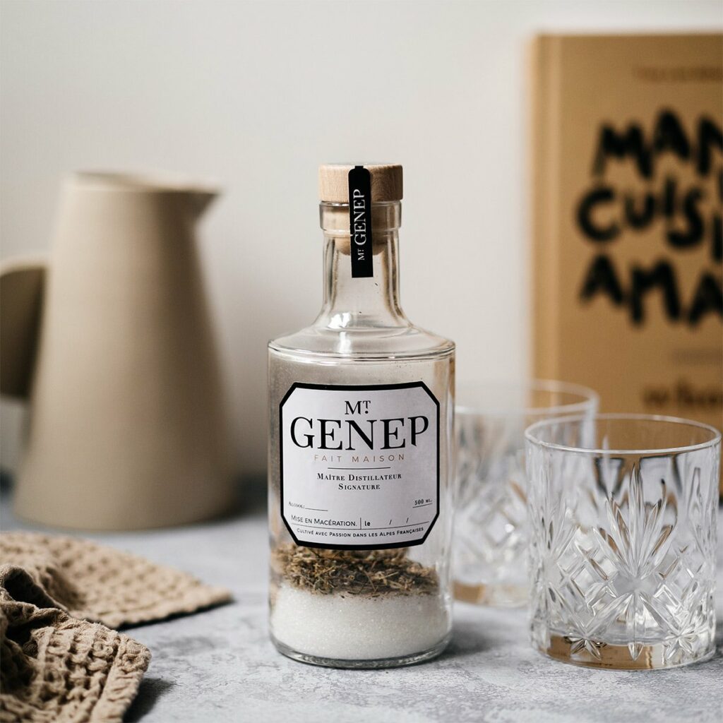Idée cadeau anniversaire DIY pour sa femme, son mari ou un proche créatif : kit de préparation de liqueur de génépi Les Raffineurs avec bouteille en verre, mélange de plantes séchées et accessoires, posé sur une table rustique avec un verre de liqueur dorée, ambiance conviviale et artisanale rappelant la montagne et les traditions fait maison.