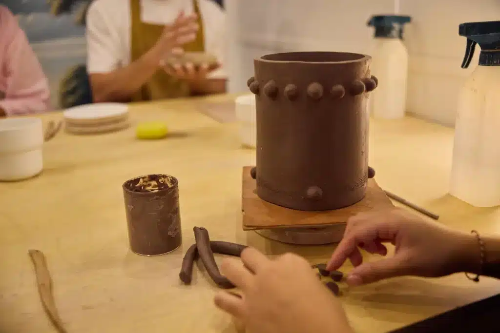 Idée cadeau anniversaire DIY pour sa femme, sa sœur ou un proche créatif : atelier poterie Wecandoo pour fabriquer ses pots en terre cuite, mains dans l’argile, outils de modelage et plantes vertes en arrière-plan, ambiance artisanale et chaleureuse mettant en valeur la création fait main et l’amour des plantes.