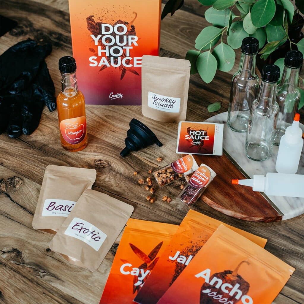 Idée cadeau anniversaire DIY pour sa femme, son mari ou un proche fan de cuisine : kit Les Raffineurs pour préparer sa sauce piquante maison, comprenant épices, bouteilles en verre et accessoires, posé sur une table en bois avec piments rouges et bols d’ingrédients, ambiance gourmande et épicée, mettant en valeur l’esprit fait main et le plaisir d’expérimenter en cuisine.