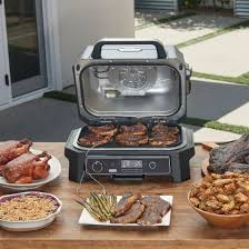 Image du barbecue électrique et fumoir Ninja Woodfire, un appareil 7-en-1 permettant de griller, fumer, rôtir, cuire au four, frire à l'air, déshydrater et réchauffer. Utilisant des granulés de bois pour une saveur authentique de fumée, il offre une expérience de cuisson en plein air polyvalente et pratique. Son design robuste et compact est idéal pour les amateurs de barbecue cherchant à diversifier leurs méthodes de cuisson. 