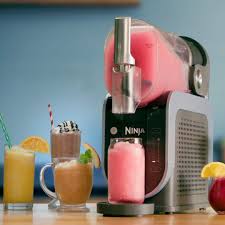 Photographie de la machine à boissons glacées Ninja SLUSHi FS301EU, un appareil au design moderne bleu arctique et gris, équipé d'un réservoir de 2,5 litres et d'un bec verseur ergonomique. Cette machine utilise la technologie RapidChill pour transformer diverses boissons en granités, milkshakes, frappés ou cocktails glacés en seulement 15 à 60 minutes, sans ajout de glace. Dotée de cinq programmes préréglés, elle permet de personnaliser la texture des boissons. Facile à utiliser et à nettoyer, avec des pièces compatibles lave-vaisselle, elle est idéale pour préparer des boissons rafraîchissantes lors de rassemblements ou pour une utilisation quotidienne à la maison