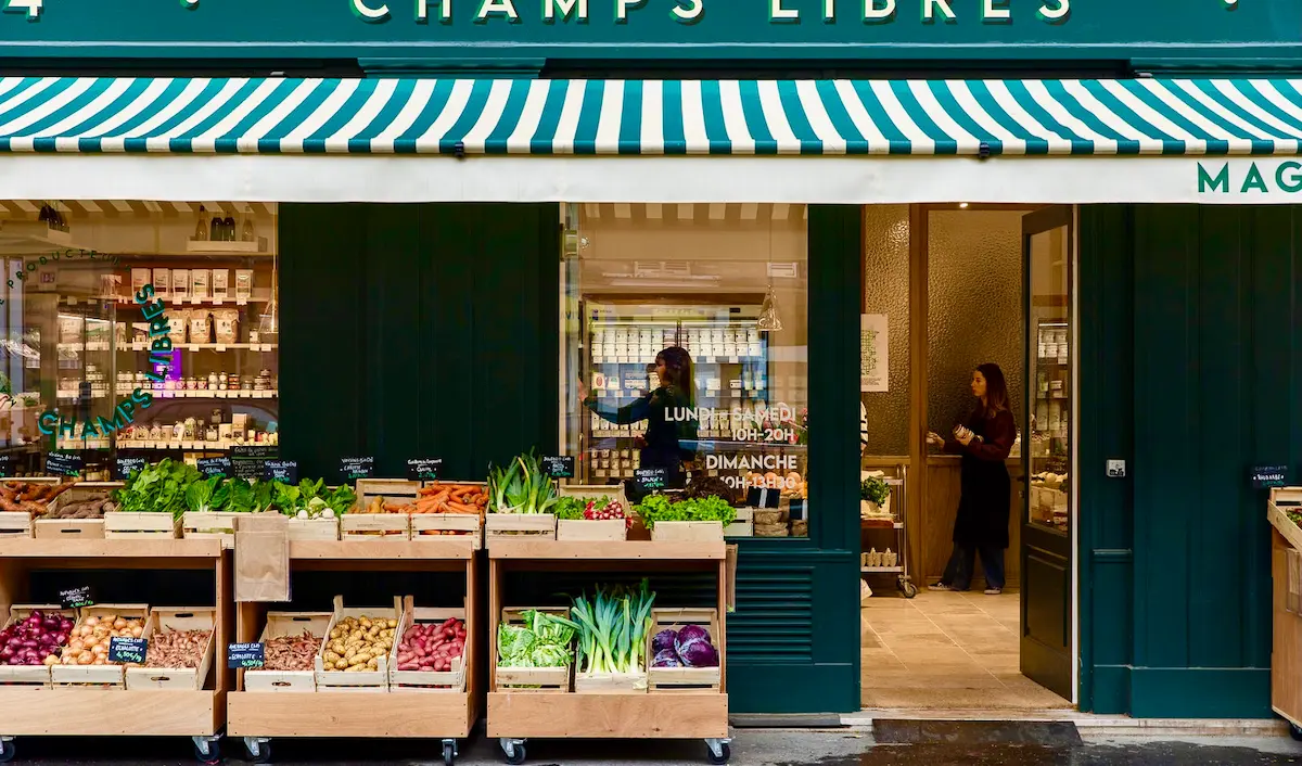 Femme consultant un panier bio sur son smartphone, comparant les prix entre un magasin bio en ligne et une boutique physique, réflexion sur comment faire ses courses bio moins cher