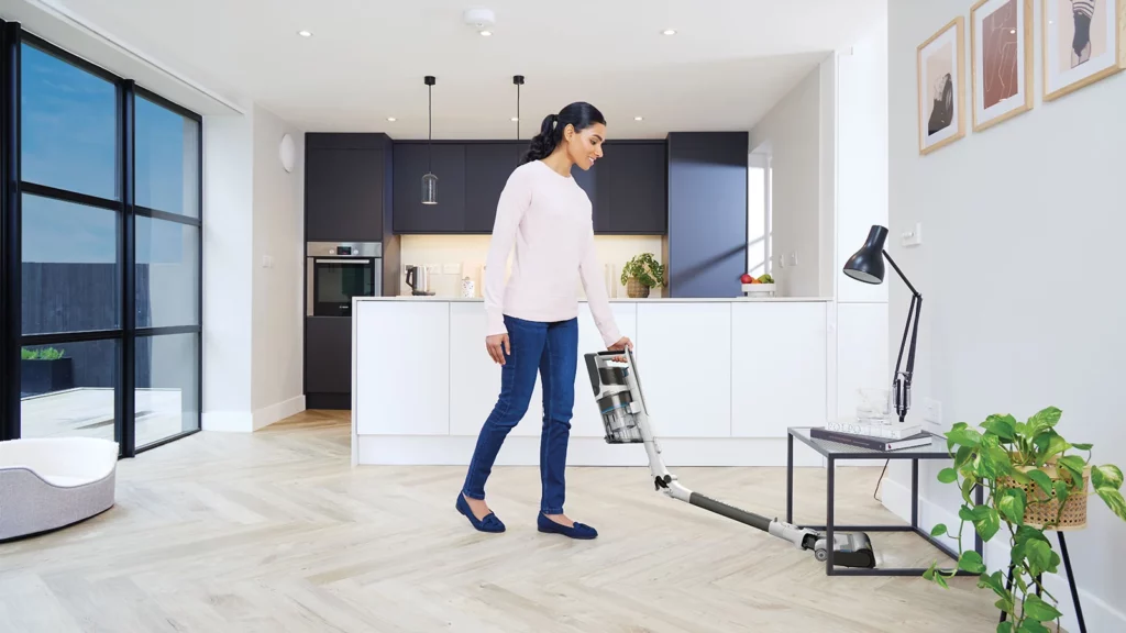 Aspirateur balai sans fil Shark PowerPro IZ380EUT en utilisation dans un intérieur avec tapis et coussins, démontrant son efficacité contre les poils d’animaux grâce à sa brosse anti-emmêlement, sa maniabilité accrue et son excellent rapport qualité-prix pour les foyers avec chiens, chats ou cheveux longs.