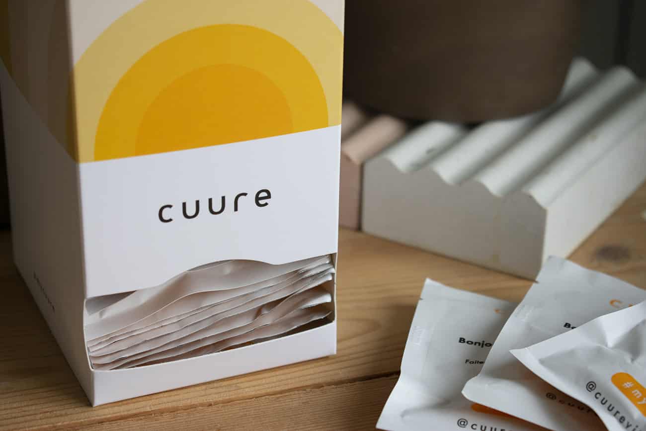 Femme assise à une table avec une boîte Cuure ouverte devant elle, découvrant les sachets journaliers personnalisés de compléments alimentaires, illustrant un avis personnel sur la marque Cuure et son service de cure bien-être à domicile, avec code promo de -20 % pour première commande.