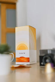 Gros plan sur la boîte Cuure fermée avec étiquette au prénom de l’utilisatrice, posée sur une table à côté d’une tasse de thé, illustrant le côté pratique, esthétique et transparent de la marque Cuure, avec avis sur le service client, l’abonnement flexible et les prix justifiés par la qualité.