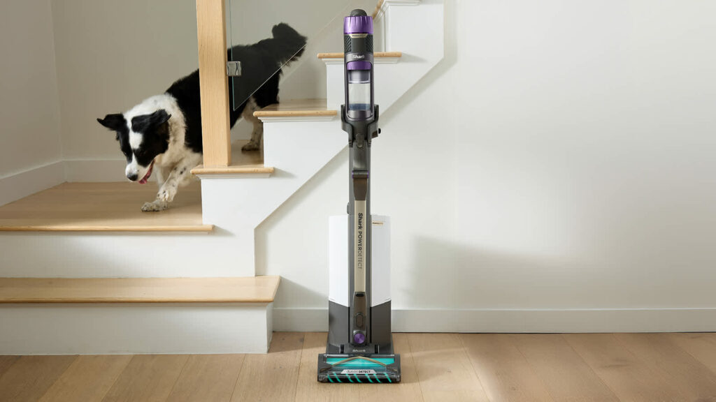 Aspirateur balai sans fil Shark PowerDetect Clean & Empty placé sur sa station d’autovidage dans un couloir lumineux, illustrant le vidage automatique du bac et la praticité du système.