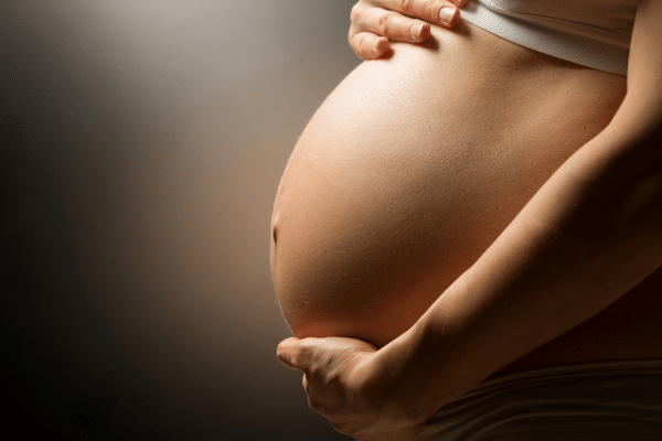 Image d’une femme enceinte tenant son ventre avec douceur, une boîte de compléments alimentaires posée sur une table de nuit, illustrant le rôle des vitamines pour la grossesse et l’allaitement
