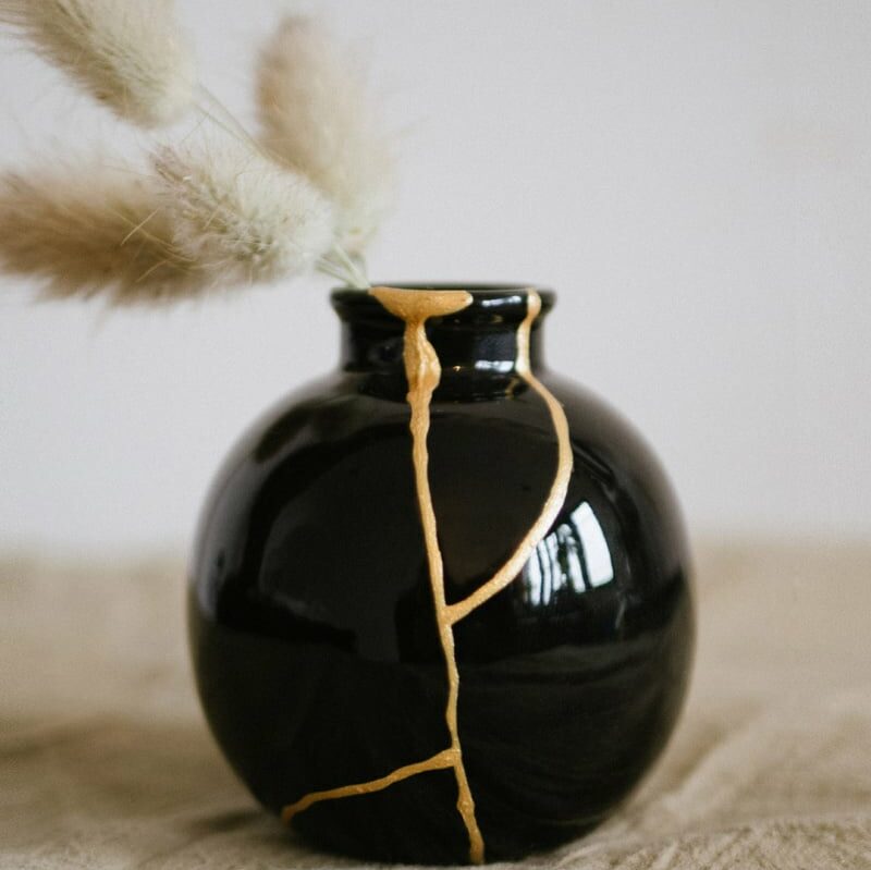 Idée cadeau anniversaire DIY pour sa femme, son épouse ou son amie créative : kit de kintsugi Les Raffineurs comprenant résine, pinceau et poudre dorée, posé sur une table en bois avec un bol en céramique réparé par des lignes dorées, ambiance artisanale et poétique inspirée du Japon, lumière douce mettant en valeur la beauté des fissures sublimées.