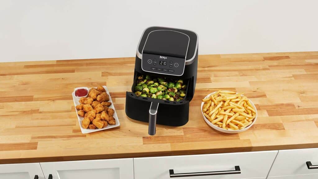 Idée cadeau d’anniversaire maison et cuisine : airfryer Ninja posé sur un plan de travail moderne avec légumes et plats préparés, ambiance lumineuse et conviviale, appareil de cuisson haut de gamme parfait pour offrir à un amateur de cuisine pratique et sain