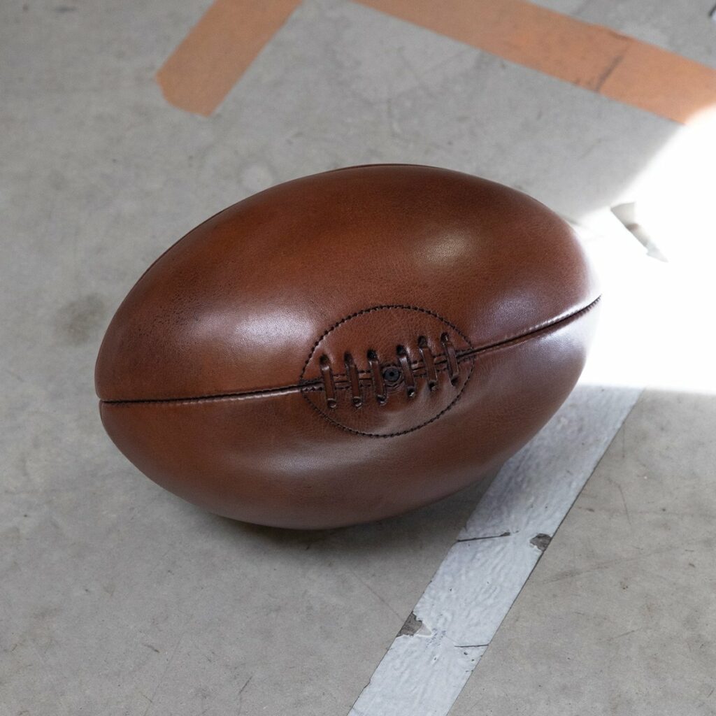 Idée cadeau anniversaire sport et déco : ballon de rugby en cuir vintage patiné, posé sur une étagère ou un banc en bois, ambiance chaleureuse et masculine, lumière douce, parfait pour un amateur de sport ou de beaux objets artisanaux.
