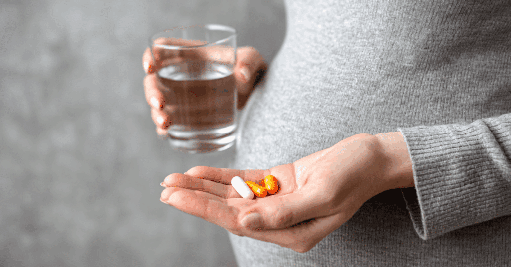 Boîte de vitamines prénatales et fruits frais, illustrant les compléments essentiels pendant la grossesse : fer, acide folique, iode et oméga 3.