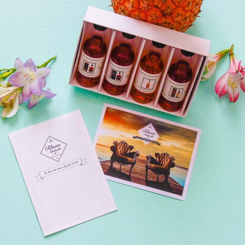 Idée cadeau anniversaire pour amateur de rhum et de spiritueux : coffret Rhum Attitude contenant plusieurs fioles de dégustation de rhums du monde, posées sur une table en bois avec verres à rhum et livret de dégustation, ambiance chaleureuse et feutrée, lumière dorée mettant en valeur les reflets ambrés des alcools, cadeau idéal pour offrir une expérience gustative raffinée à un proche.