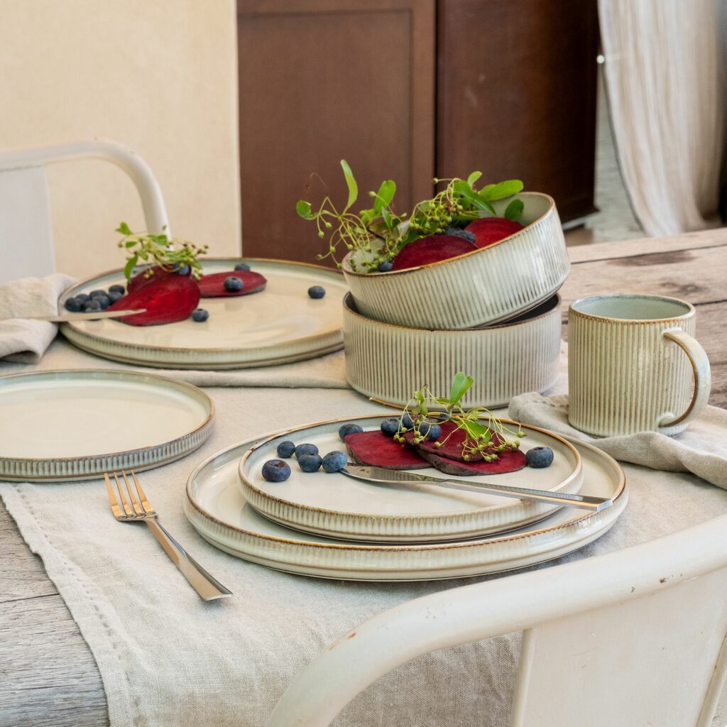 Idée cadeau d’anniversaire maison et déco : service de vaisselle en grès Alinea comprenant assiettes et bols dans des tons naturels, posé sur une table en bois, ambiance conviviale et élégante, lumière douce mettant en valeur la texture noble du grès.