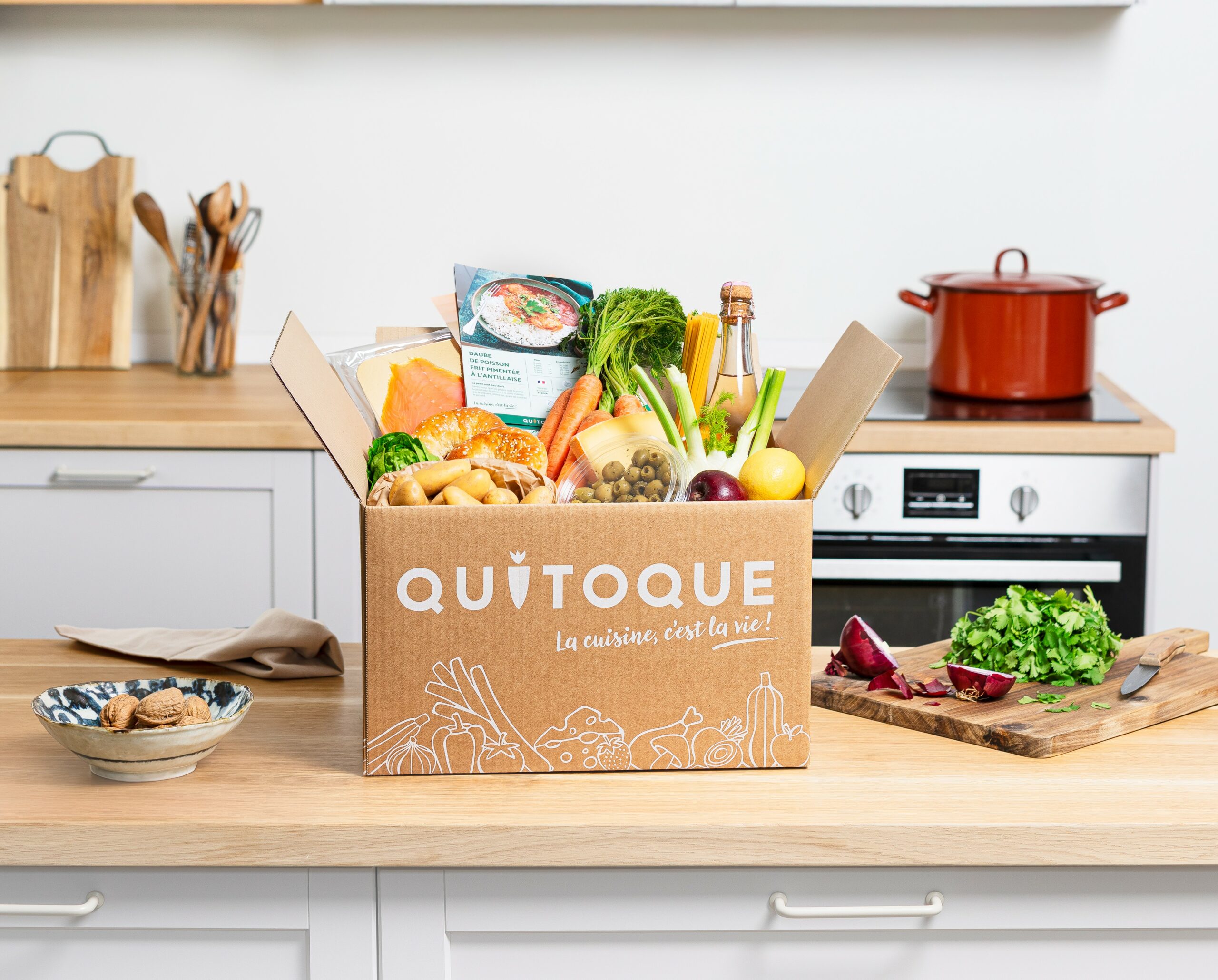 Box Quitoque ouverte sur un plan de travail de cuisine avec légumes frais, fiches recettes illustrées et paniers repas colorés. Image représentant le test et avis Quitoque France et introduisant la marque de box à cuisiner française.