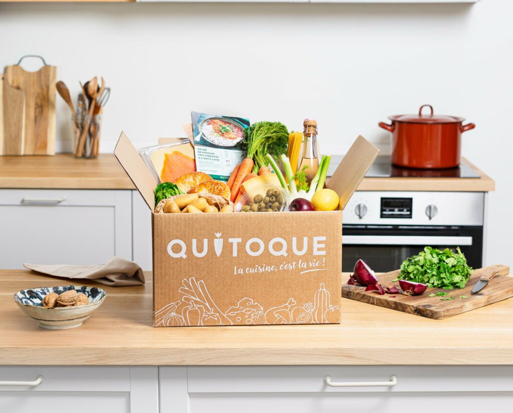 Box Quitoque ouverte avec produits frais français, légumes de saison et fiches recettes sur une table en bois rustique dans une cuisine lumineuse.