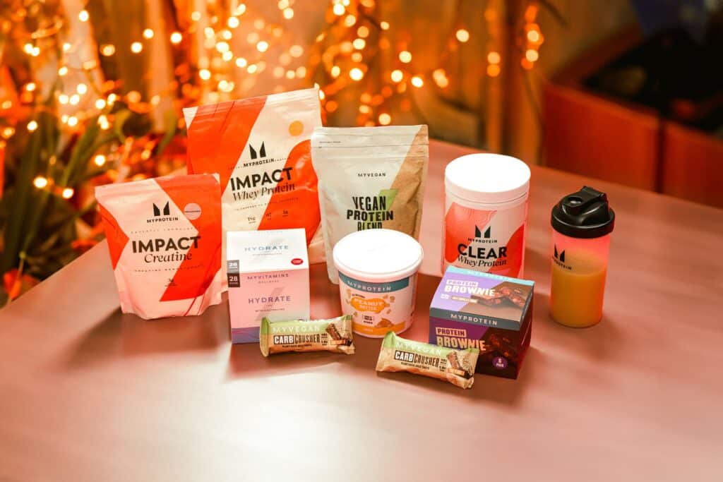 Plusieurs produits MyProtein disposés sur une table, sachet d'Impact Whey Protein, pot de créatine monohydrate et barres protéinées de la gamme, photo illustrant les meilleurs produits à commander avec un code promo MyProtein pour faire baisser la note, codes de réduction disponibles en permanence sur le site officiel fr.myprotein.com, remises allant jusqu'à 50 pourcent sur la whey et la créatine lors des ventes flash, livraison gratuite à partir de 55 euros, avis sur les produits MyProtein les plus rentables à acheter en promotion