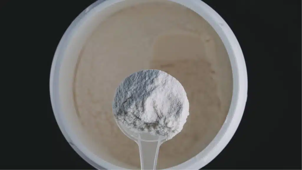 la cuillère doseuse blanche remplie de poudre de créatine, posée directement sur l’étiquette du pot pour qu’on voie en même temps la texture fine et cristalline de la poudre et le tableau de composition en arrière-plan, avec la mention du grade Creapure bien visible.